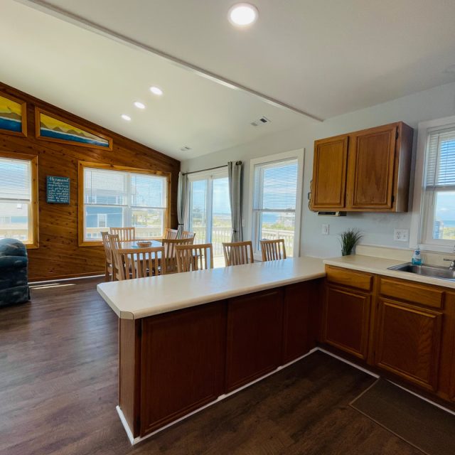 Open kitchen and dining area with wood paneling, large windows, and view of houses and water outside.