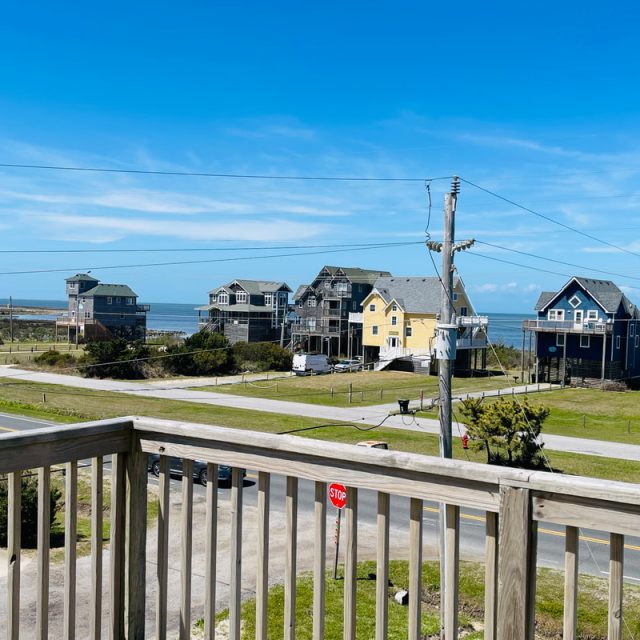 View from a wooden deck overlooking a street, beach houses, power lines, and the ocean under a clear blue sky.
