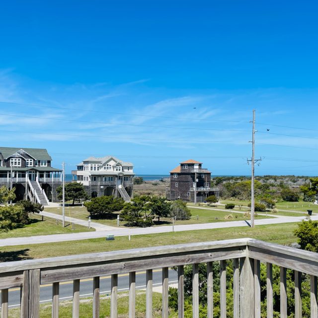 View from a wooden deck overlooking houses, grassy yards, and a distant ocean under a clear blue sky.