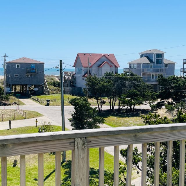 View from a wooden balcony overlooking beach houses, trees, and a clear blue sky near the ocean.