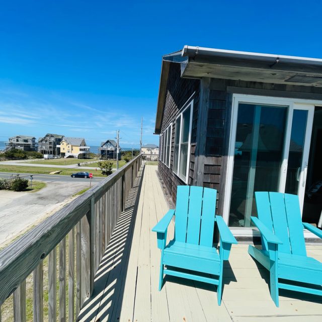 Two turquoise Adirondack chairs on a wooden deck outside a house, with a view of neighboring homes and a clear blue sky.