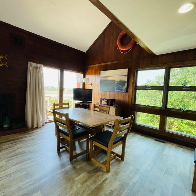 Dining area with wooden table and chairs, TV, artwork, and large windows showing greenery outside.