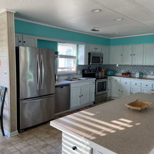 Modern kitchen with white cabinets, stainless steel appliances, and turquoise walls, lit by natural sunlight.