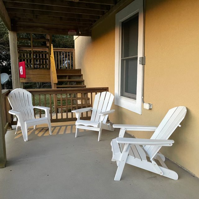 Three white Adirondack chairs are arranged on a covered porch next to a yellow building with a window.