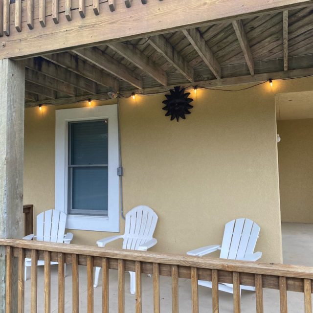 Three white Adirondack chairs sit on a covered porch with string lights and a sun-shaped wall decoration.