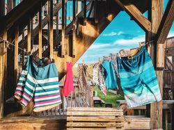 Swimwear and towels hang on a clothesline under wooden stairs with a bright blue sky in the background.