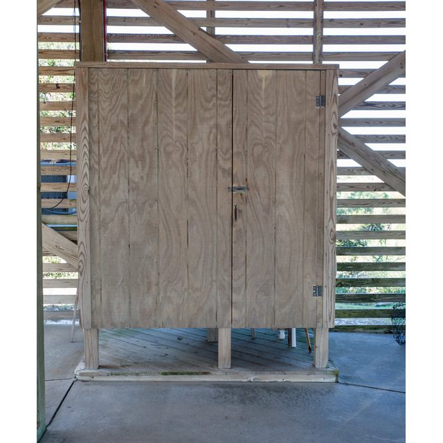 A wooden cabinet with double doors and a latch stands on a concrete floor in front of a slatted wooden wall.