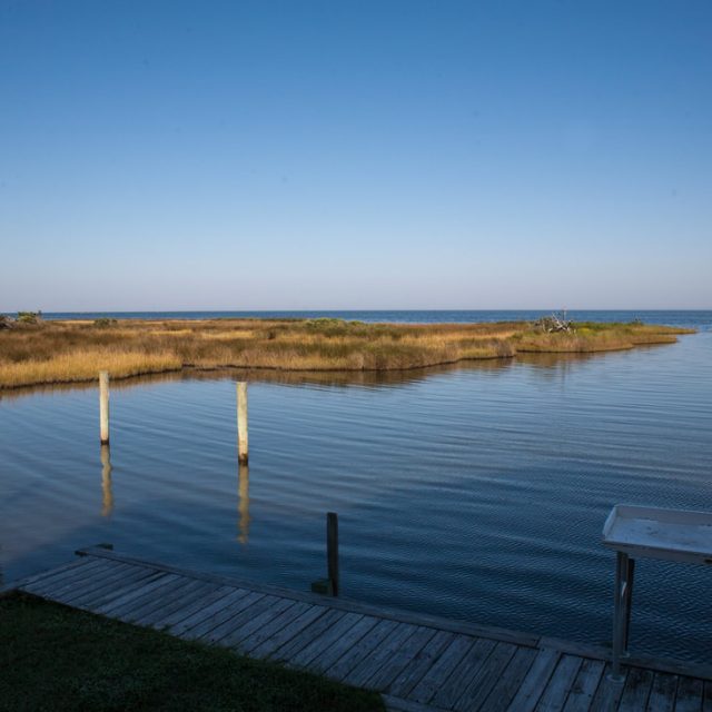 Wooden dock extends over calm water with marsh grasses and clear blue sky in the background.