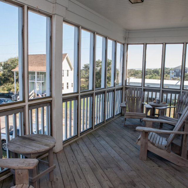 Screened porch with wooden chairs and tables, overlooking houses and greenery on a sunny day.