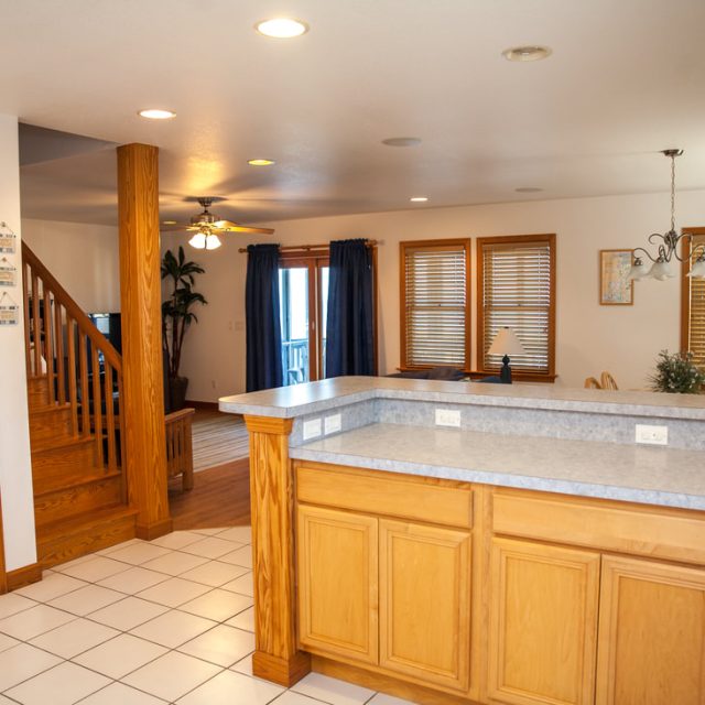 Open kitchen with light wood cabinets, white tile floor, and a view of the dining and living areas with wooden staircase.