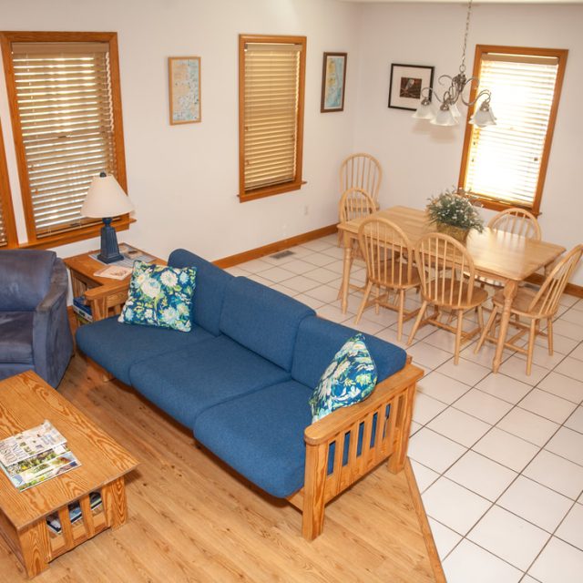 Living room with blue sofa, armchair, wooden table, dining area, tile floor, and three windows with blinds.