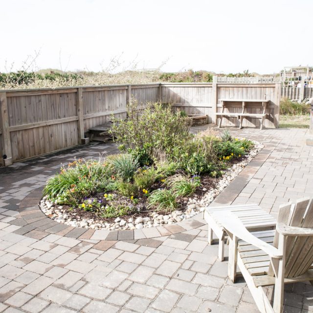 Brick patio with wooden chairs, a bench, and a small landscaped garden bed surrounded by a wooden fence.