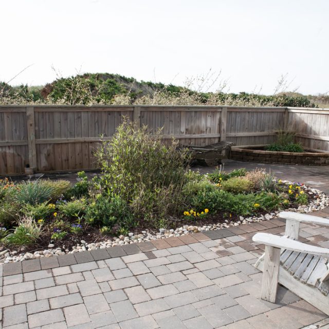 A fenced patio with paver stones, a central garden bed, wooden benches, and a white wooden chair.
