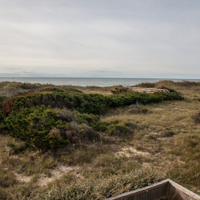 A sandy, grassy dune landscape leads to the ocean under a cloudy sky, with a wooden walkway in the distance.