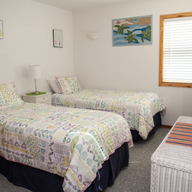 Bedroom with two twin beds, pastel bedding, a window with blinds, wicker furniture, and colorful wall art.