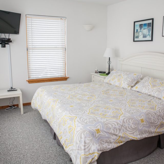 Bedroom with a large bed, patterned bedding, wall-mounted TV, two windows, and framed photos above the headboard.
