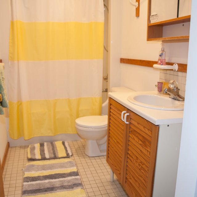 Bathroom with a yellow and white shower curtain, striped rug, toilet, sink, and wooden cabinets.