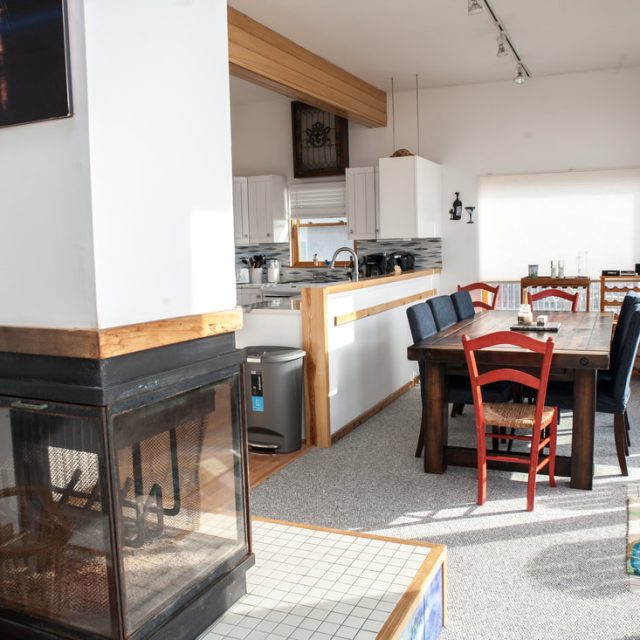 Open kitchen and dining area with a wooden table, mismatched chairs, fireplace, and large windows letting in sunlight.