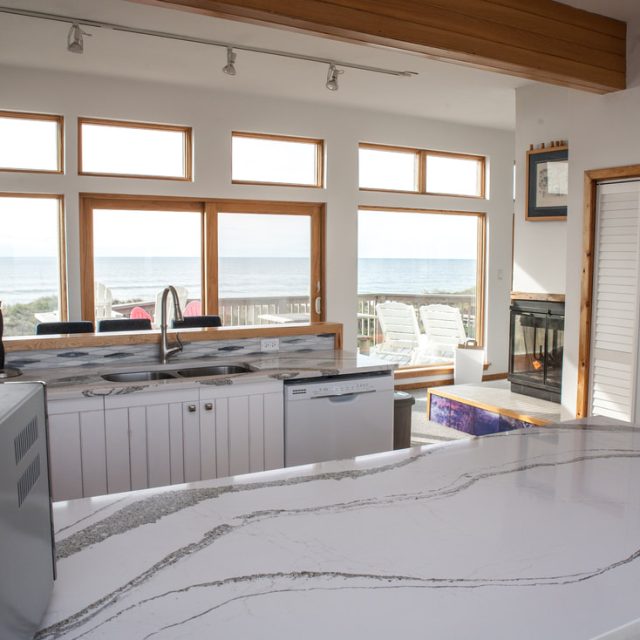 Modern kitchen with white cabinets, marble countertop, large windows, and an ocean view in the background.