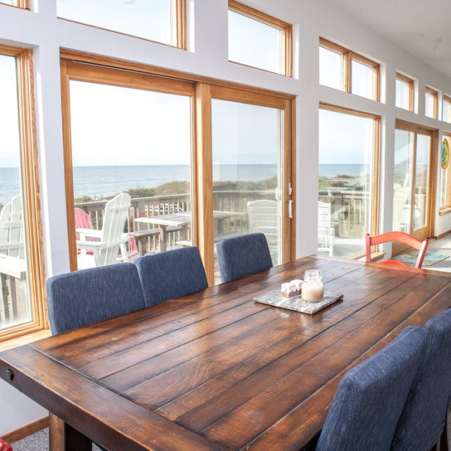 Wood dining table and blue chairs in a bright room with large windows overlooking a beach and ocean view.