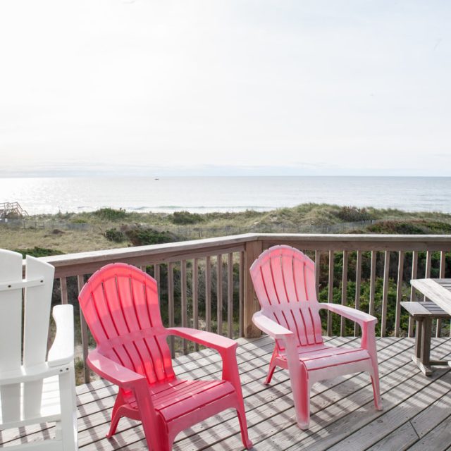 Three empty chairs and a wooden table on a deck overlook grassy dunes and a calm ocean under a bright sky.