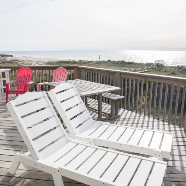 Two white lounge chairs and a picnic table on a wooden deck overlooking a beach and ocean under a bright sky.
