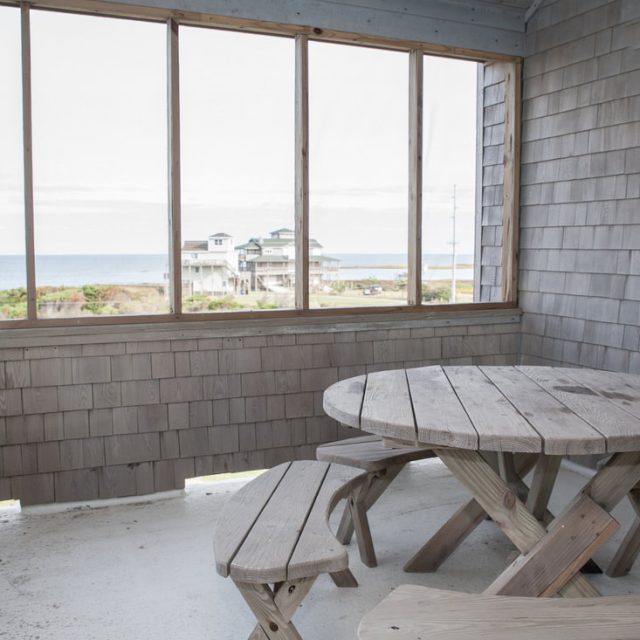 Covered patio with a round wooden picnic table and benches, overlooking the ocean and nearby houses through screened windows.