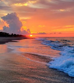 Waves roll onto a sandy beach at sunset, with clouds and vibrant orange and pink colors in the sky.