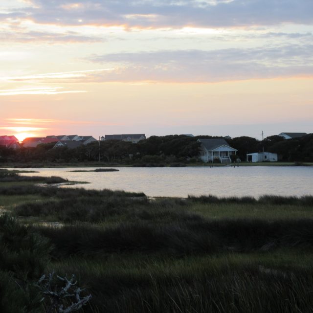 Sunset over a row of houses by a calm lake, with marsh grasses in the foreground and clouds in the sky.