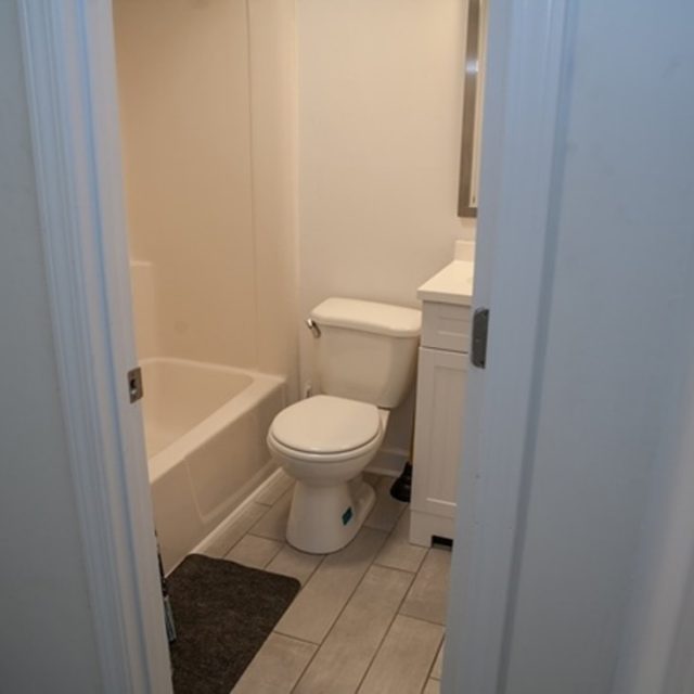 Small bathroom with a toilet, white vanity, bathtub, gray floor tiles, and an open door.