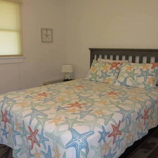 Bedroom with a bed covered in a starfish-patterned bedspread, a nightstand, lamp, and window with closed blinds.