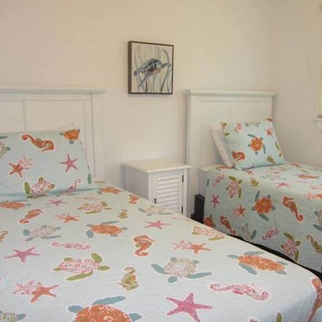 Two twin beds with sea-themed bedding in a simple room with a nightstand, wall art, and a window with blinds.