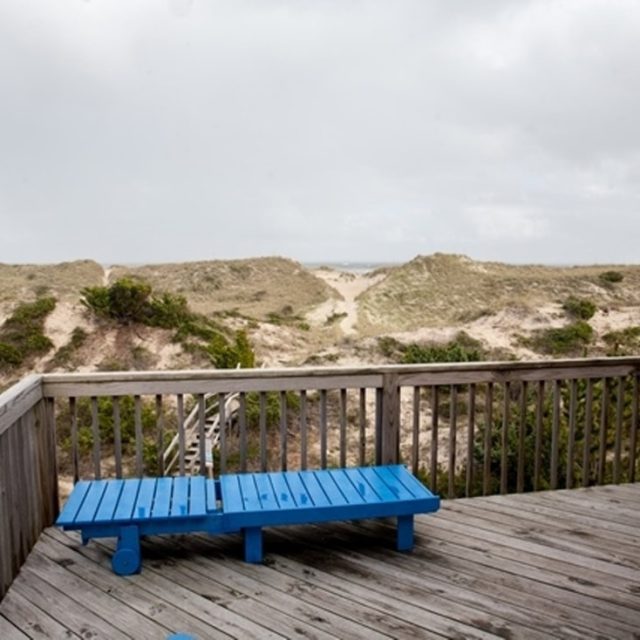 Wooden deck with two blue lounge chairs overlooks sandy dunes under a cloudy sky.