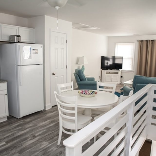 Open-plan kitchen and living room with white furniture, wood floors, and blue armchairs near a TV.