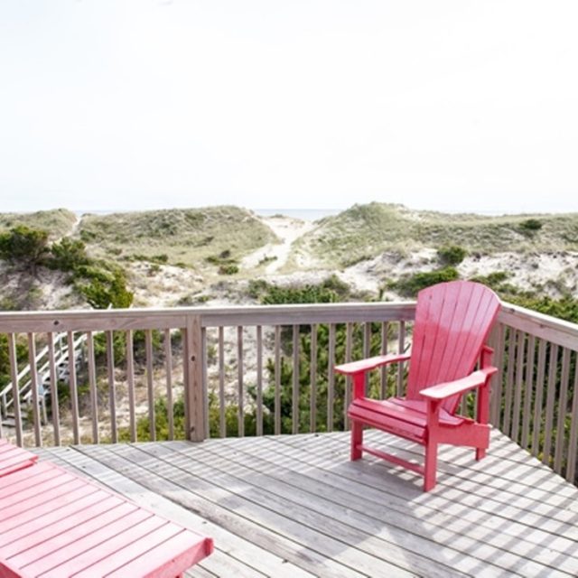 Wooden deck with red Adirondack chairs overlooks sandy dunes and greenery under a bright, clear sky.